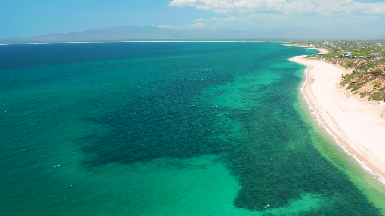 Aerial frame over El Sargento La Ventana, Mexico, with turquoise water in the foreground, darker underwater patches, a pale beach at right, distant shoreline, and tiny distant people near the surf.