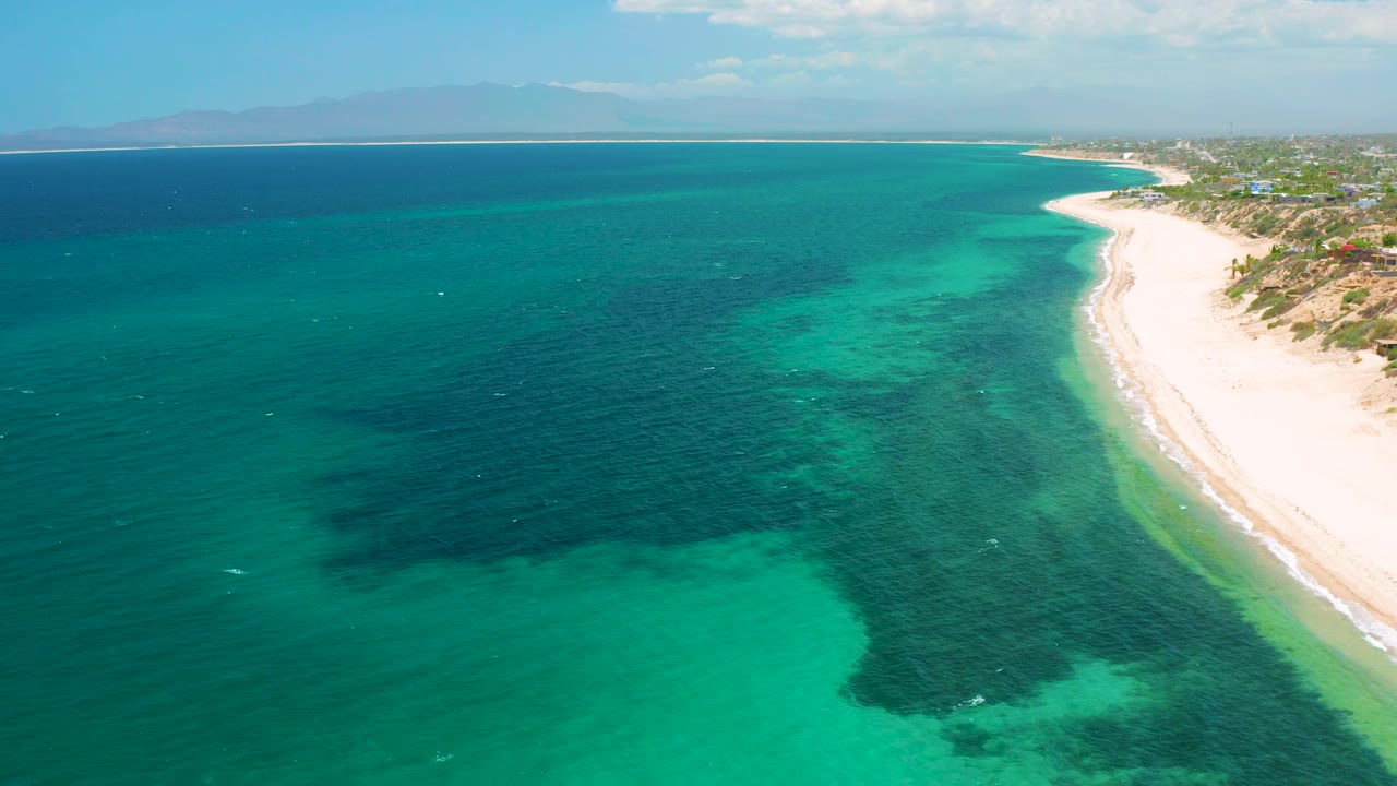 Wide aerial view in El Sargento La Ventana, Mexico, showing darker reef or seaweed shapes under clear turquoise water, the beach compressed along the right side, and no close identifiable people.