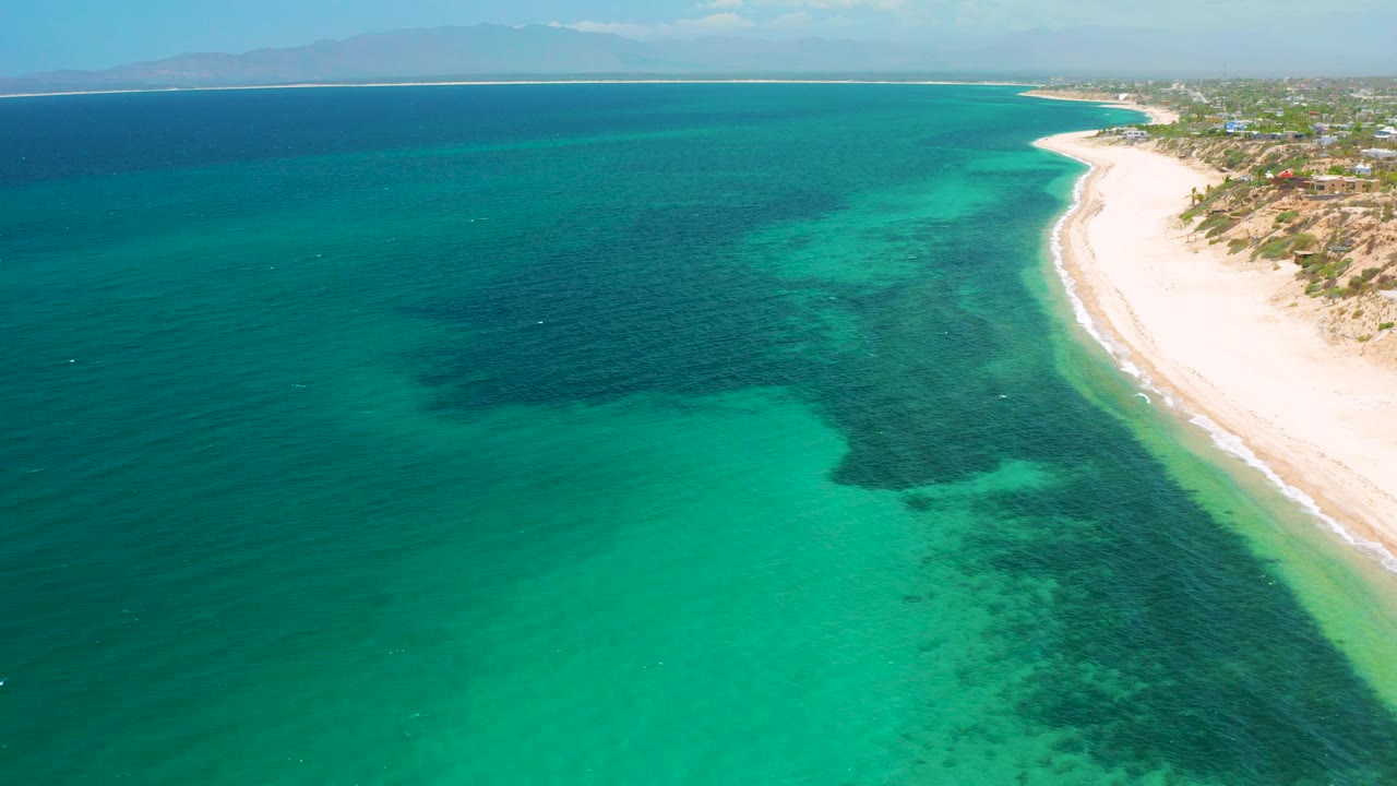 Aerial frame over El Sargento La Ventana, Mexico, with turquoise shallows in the foreground, darker underwater patches, a pale beach on the right, distant land on the horizon, and no visible people.