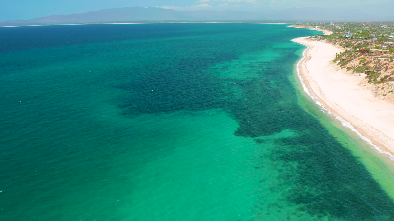 Drone view over El Sargento La Ventana, Mexico, with emerald water filling most of the frame, a dark submerged patch near center, bright sand along the right, coastal buildings beyond, and no visible people.
