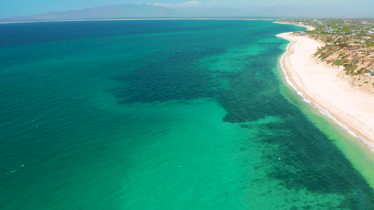 High aerial frame over El Sargento La Ventana, Mexico, showing clear blue-green water, a broad dark underwater area, a narrow bright beach at right, low development near shore, and no visible people.