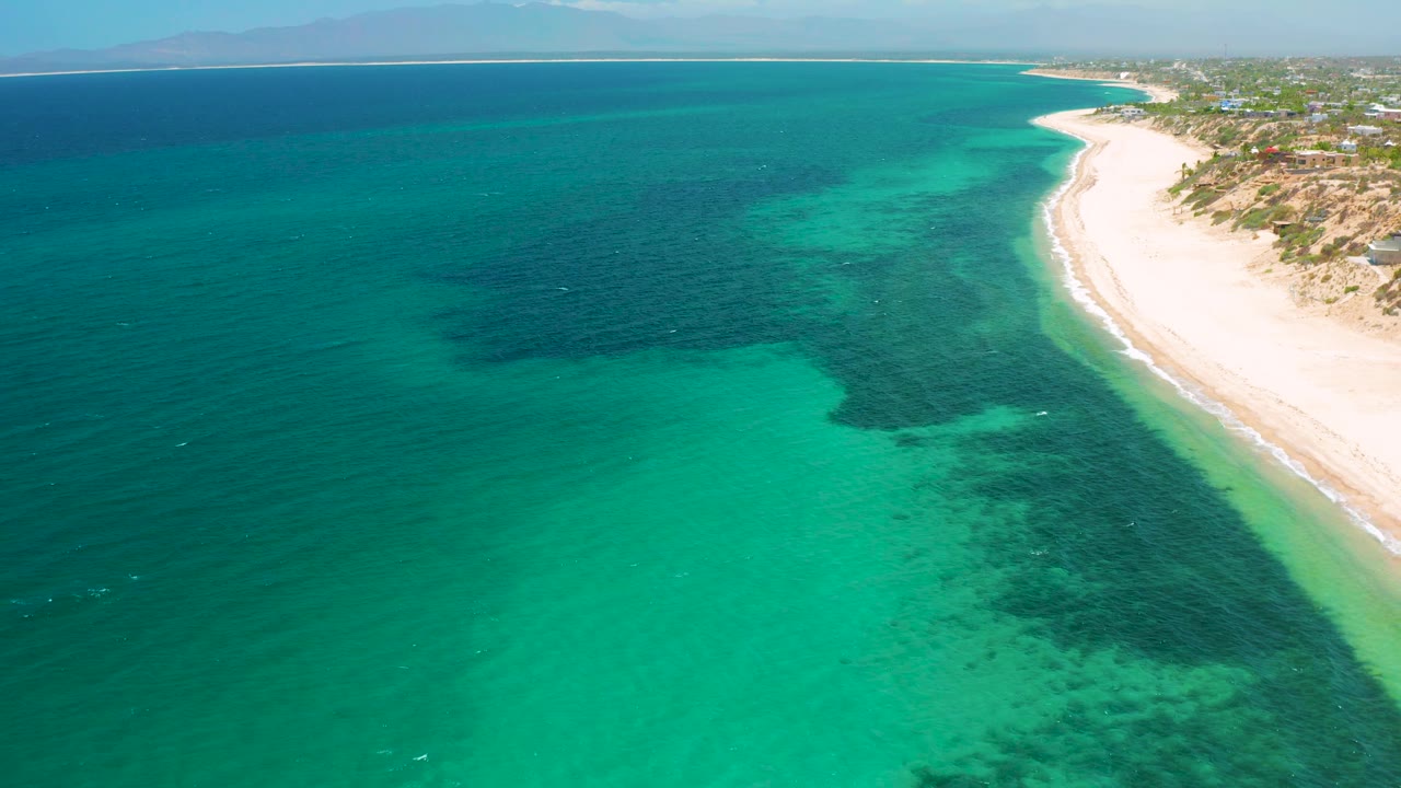 Aerial view in El Sargento La Ventana, Mexico, with green water in the foreground, dark reef-like or seaweed patches below the surface, a pale beach strip to the right, distant mountains or landforms, and no visible people.