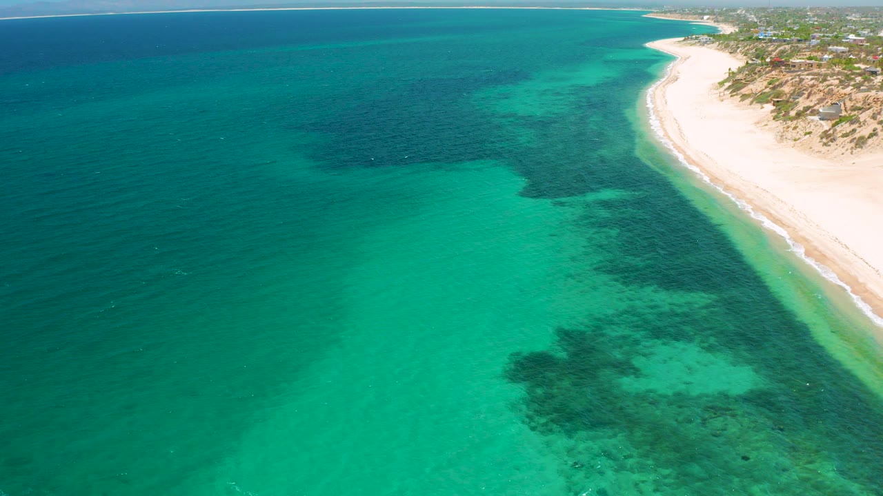 Aerial view over turquoise water and dark underwater patches beside the curving beach at El Sargento La Ventana, Mexico, with no visible people.