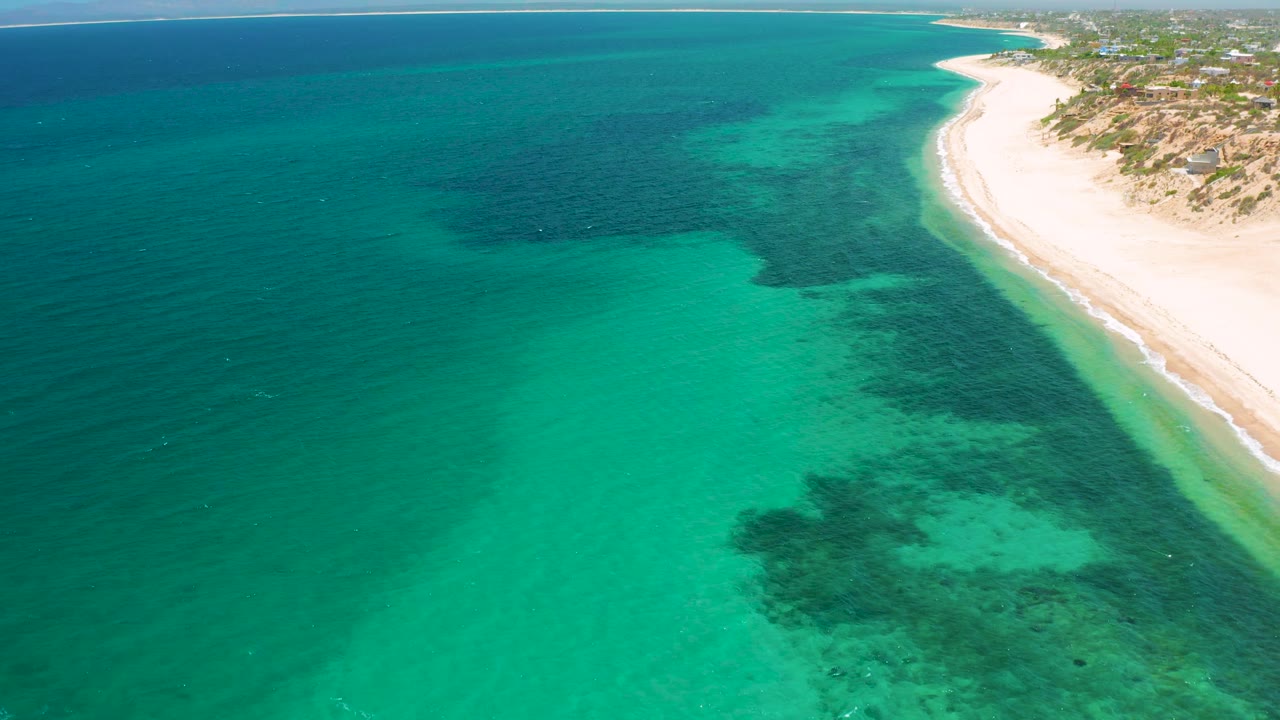 Drone view of aqua sea, darker submerged areas, and a bright sand shoreline at El Sargento La Ventana, Mexico, with no visible people.