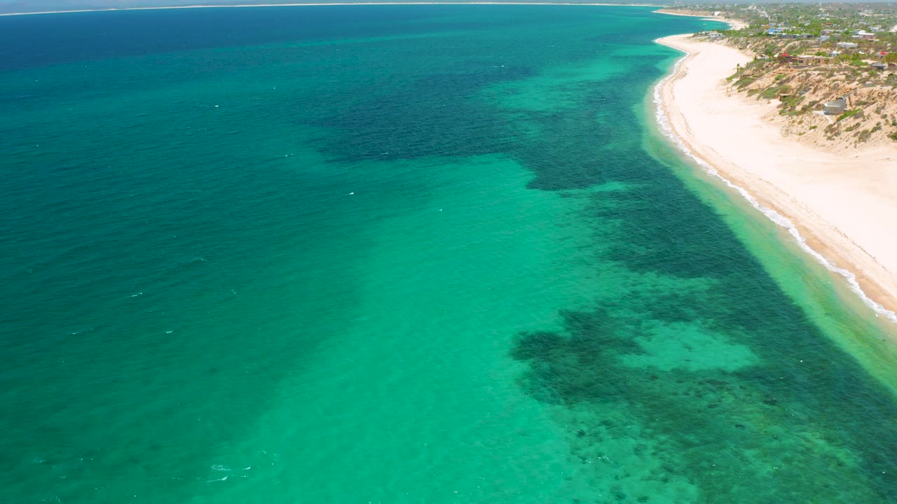 High aerial frame of turquoise shallows, dark reef or sea grass shapes, and the sandy coast of El Sargento La Ventana, Mexico, with no visible people.