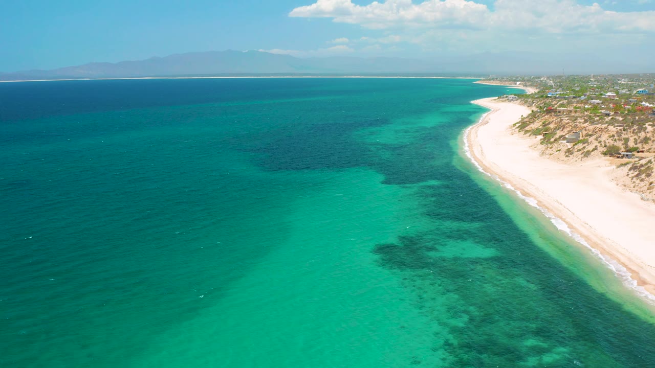 Aerial frame from El Sargento La Ventana, Mexico, showing broad turquoise shallows, dark underwater patches, a narrow strip of white sand at right, and no visible people.