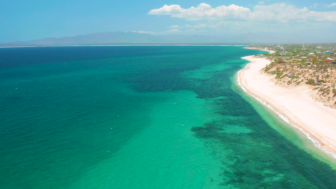 Aerial frame from El Sargento La Ventana, Mexico, with open water across the left side, pale sand and shrubs along the right, distant mountains, and no visible people.