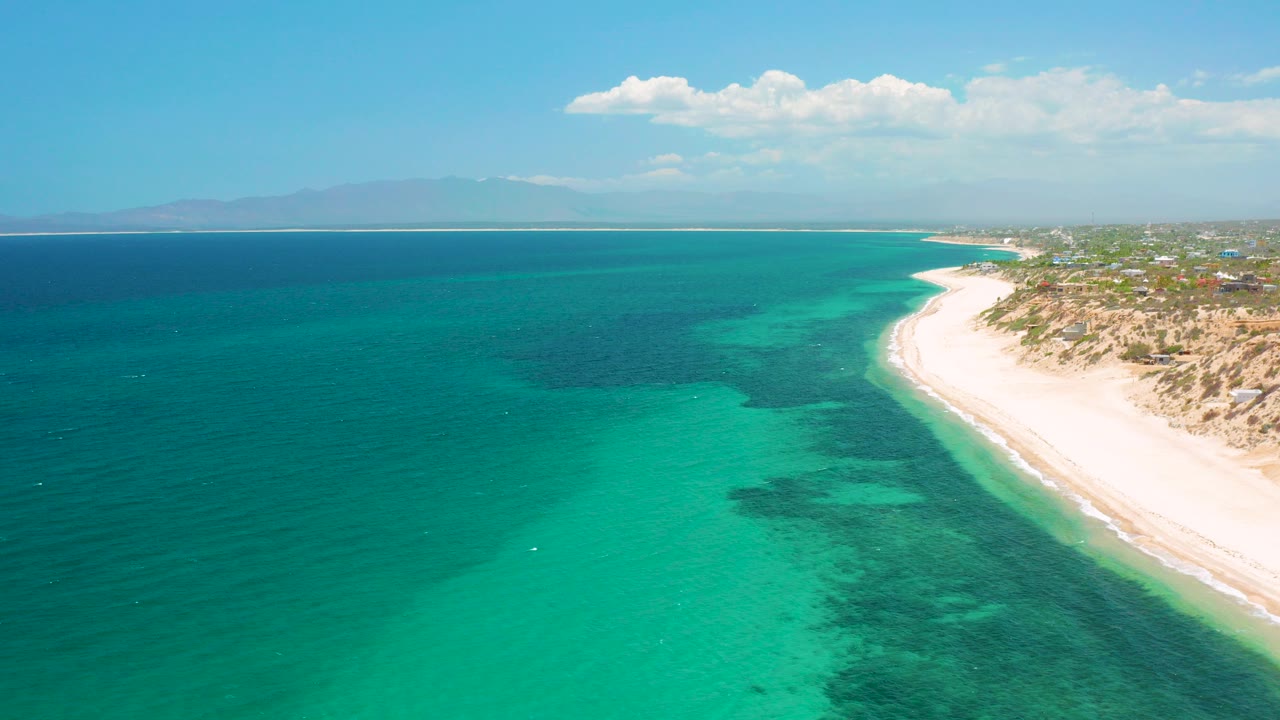 Aerial view over El Sargento La Ventana, Mexico with turquoise sea, a pale beach curving along the right edge, distant mountains, scattered clouds, and no visible people.