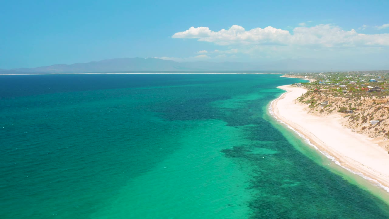 Aerial view in El Sargento La Ventana, Mexico with turquoise water in the foreground, darker underwater patches, white beach at right, hazy mountains on the horizon, and no visible people.