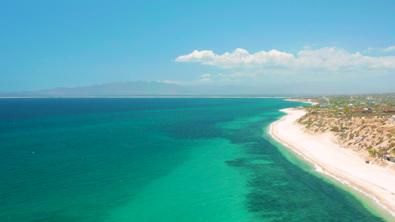 Opening aerial frame over El Sargento La Ventana, Mexico, with turquoise water in the foreground, a pale beach curving along the right edge, distant mountains under scattered clouds, and no visible people.