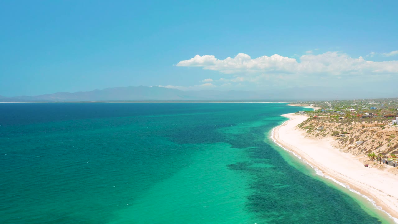 Aerial frame over El Sargento La Ventana, Mexico, showing clear green-blue shallows, darker underwater patches, a bright sand strip to the right, distant coastline ahead, and no visible people.