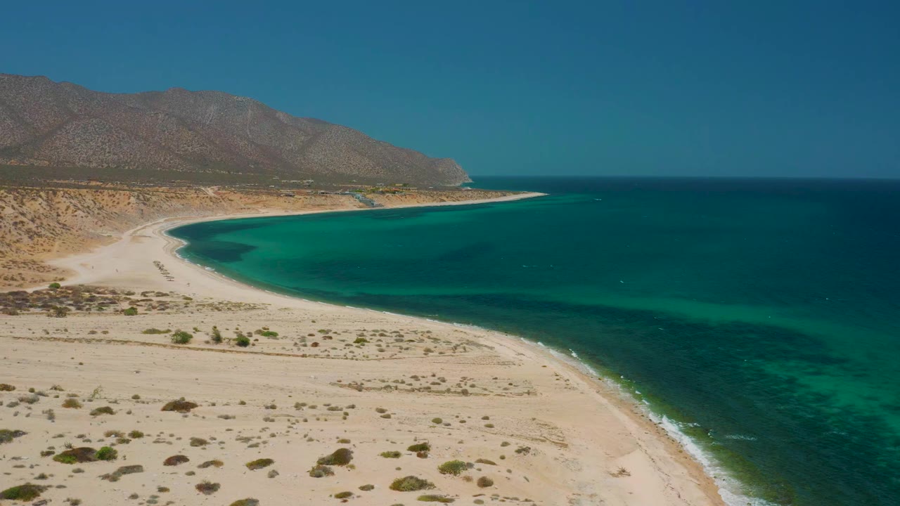 Wide aerial view in El Sargento La Ventana, Mexico, showing the shoreline wrapping around blue-green water with dry beach terrain below, mountains on the left horizon, and no visible people.