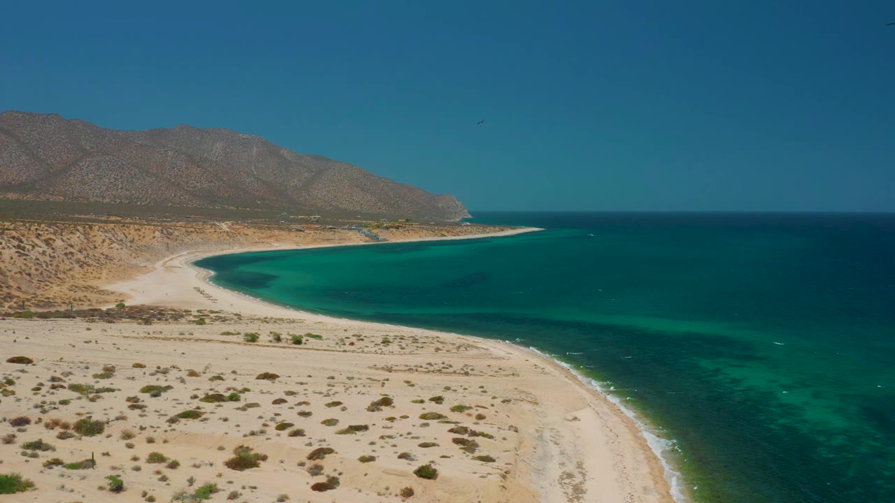 At 0:01, the aerial view holds on the El Sargento La Ventana shoreline, with scrubby sand in the lower frame, a bright beach arc, green-blue water, distant hills, and no visible people.