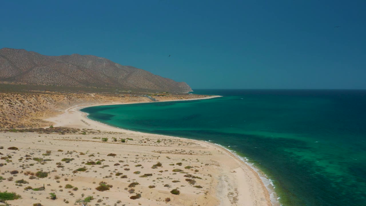 At 0:02, El Sargento La Ventana is seen from above with the beach bend centered, clear green water along the shore, deeper blue sea beyond, mountain slopes to the left, and no visible people.