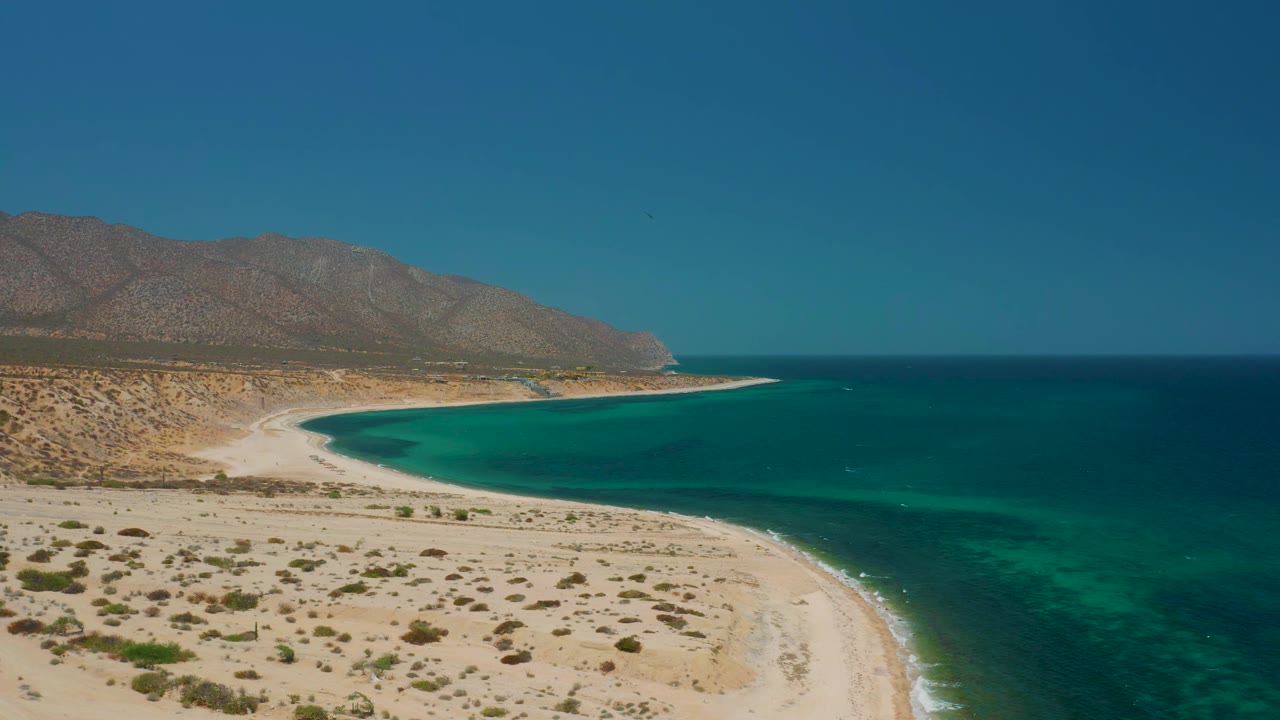 A high aerial view in El Sargento La Ventana, Mexico, with desert scrub on pale sand, blue-green water along the right side, distant mountains, and no visible people.