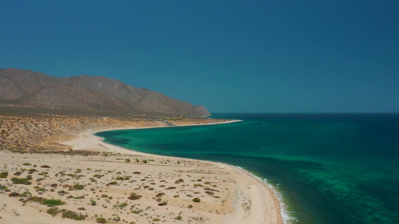 Wide aerial coastline at El Sargento La Ventana, Mexico, where an empty sandy foreground leads to a clear bay, darker offshore water, mountain slopes, and no visible people.
