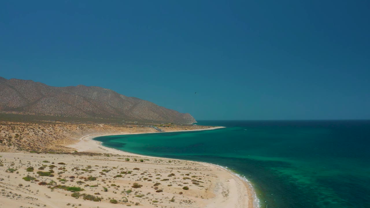 Aerial frame over El Sargento La Ventana, Mexico showing turquoise shallows along a pale sandy beach, desert shrubs in the foreground, a brown mountain ridge, and no visible people.