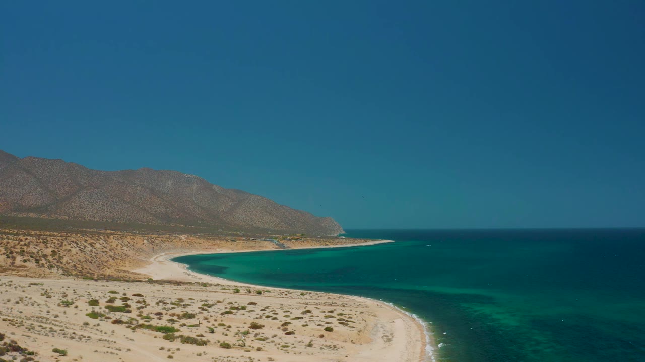 A wide aerial moment over El Sargento La Ventana, Mexico, showing the turquoise bay filling the center, sandy beach at lower left, mountain ridge behind it, and no visible people.