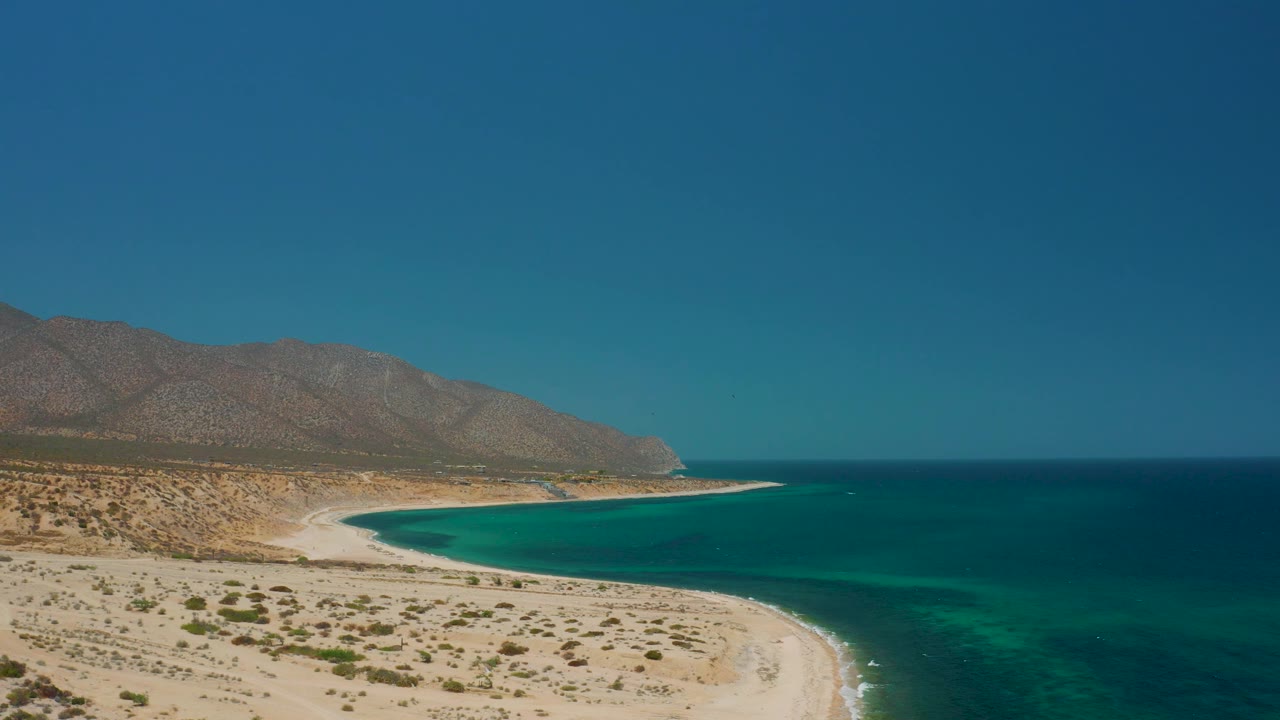 A later aerial frame of El Sargento La Ventana, Mexico, with the sandy crescent beach running below the mountains, darker offshore water to the right, and no visible people.