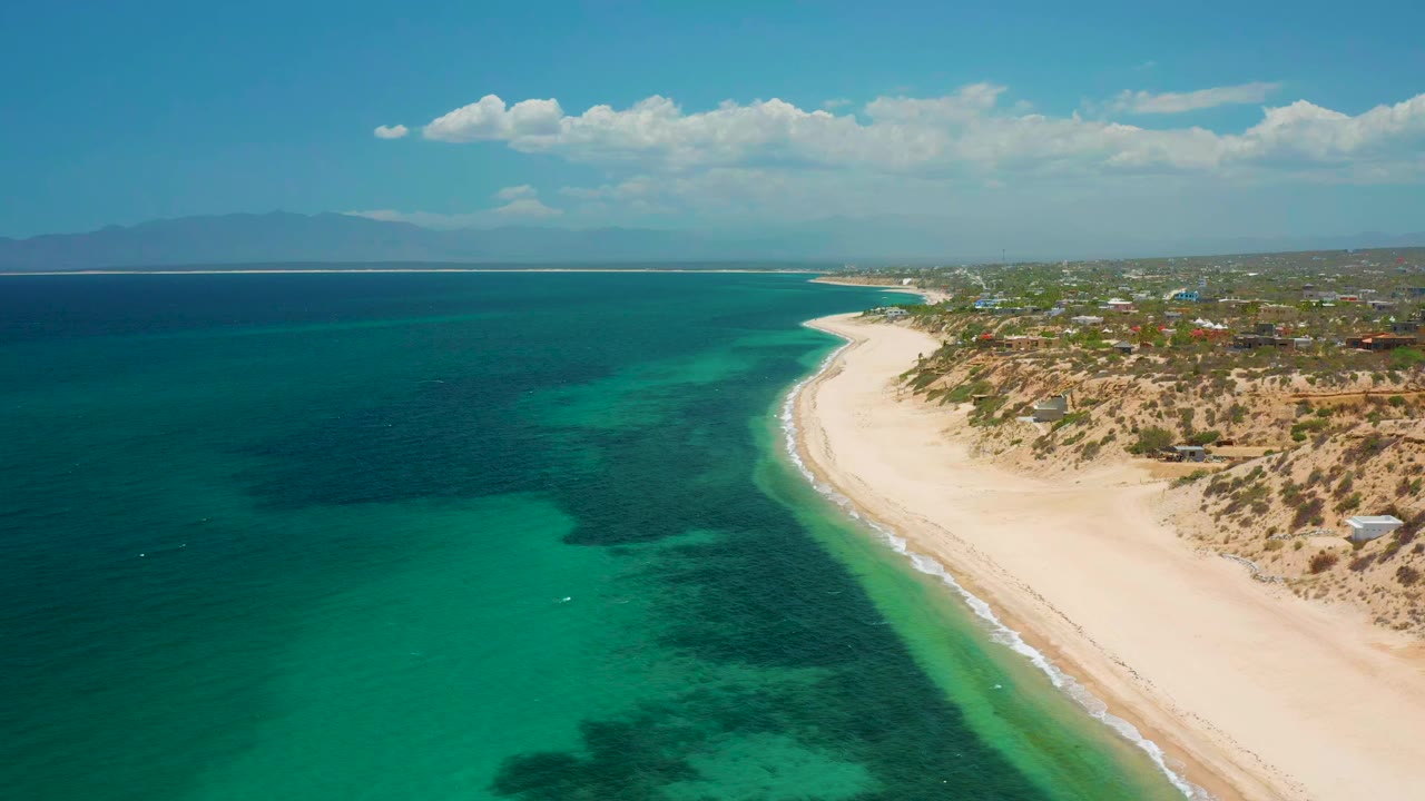 Aerial frame over El Sargento La Ventana, Mexico, with turquoise water on the left, a curving sandy beach on the right, distant mountains on the horizon, and no visible people.
