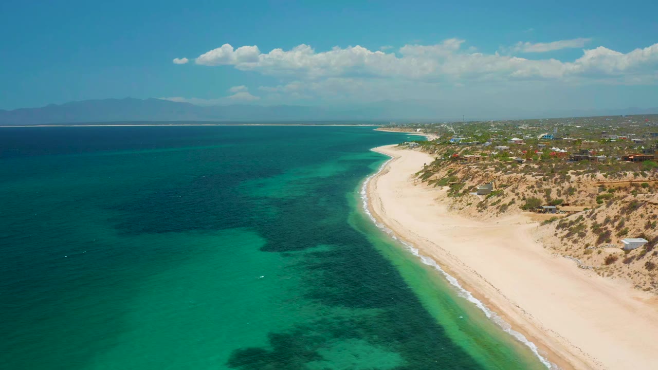 Aerial view of the El Sargento La Ventana shoreline with deeper blue water offshore, lighter turquoise water near the beach, low buildings inland, and no visible people.