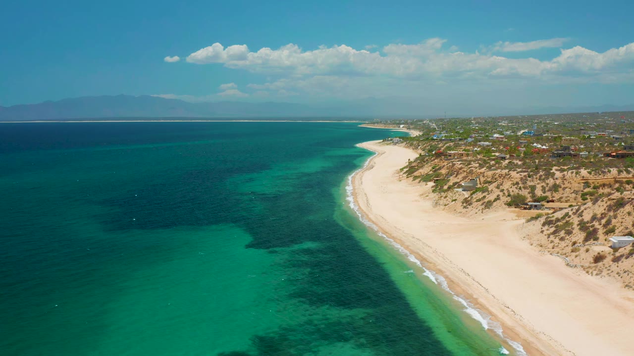 High coastal view with clear water, sandy shoreline, and buildings beyond dunes.