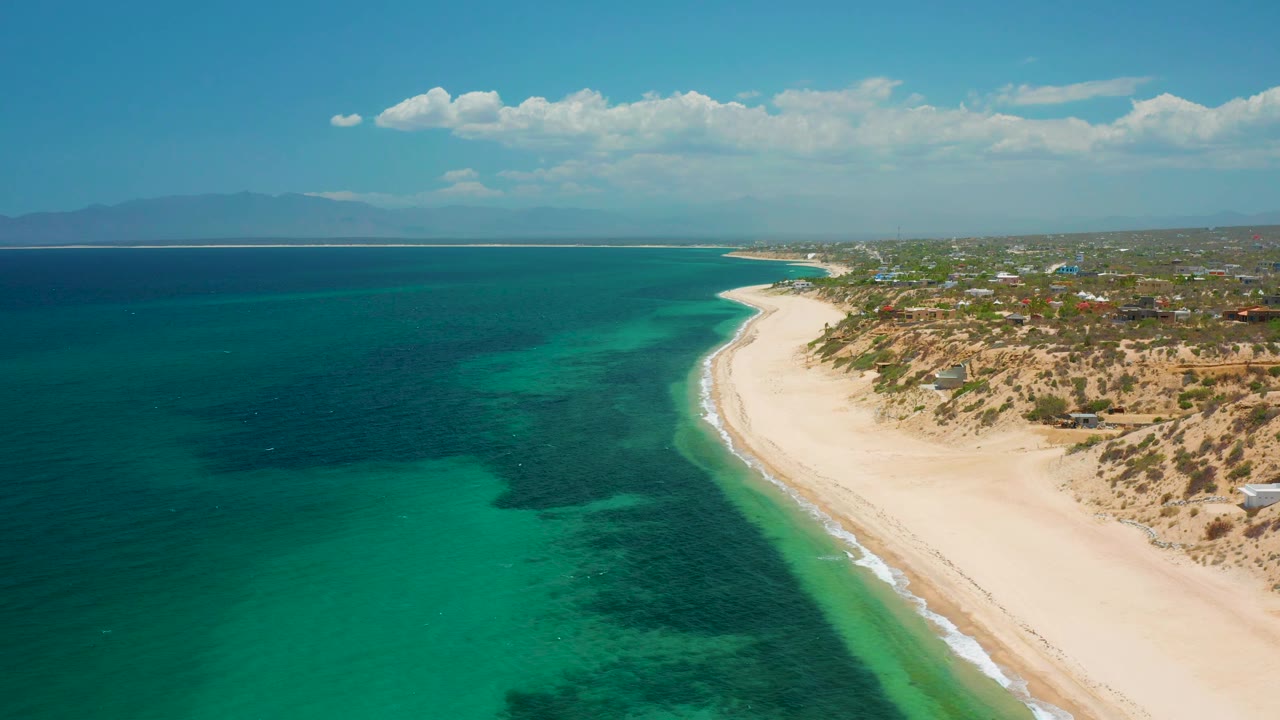 The aerial frame shows El Sargento La Ventana coastline with dark underwater patches near turquoise shallows, a broad pale beach, dry vegetated dunes, inland rooftops, and no visible people.