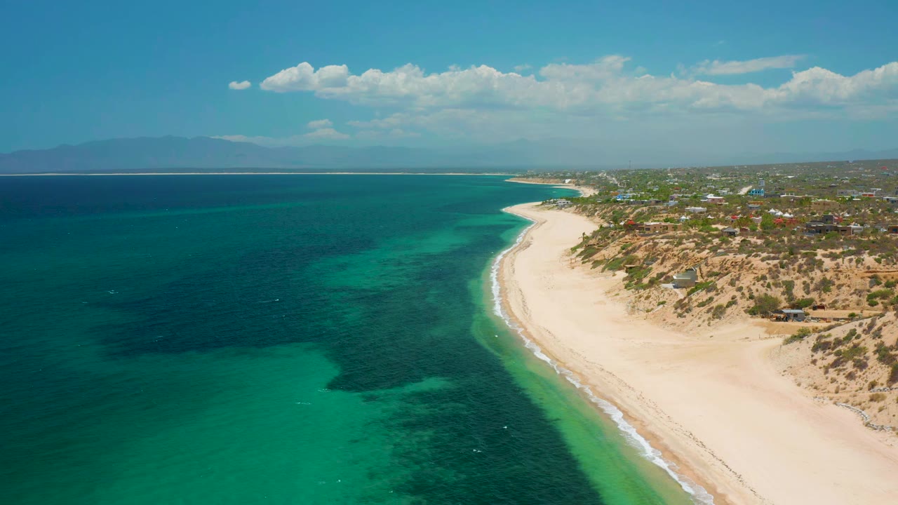 High drone view of El Sargento La Ventana, Mexico, showing blue-green shallows, darker offshore water, a long pale beach, scattered shoreline buildings, distant horizon, and no visible people.