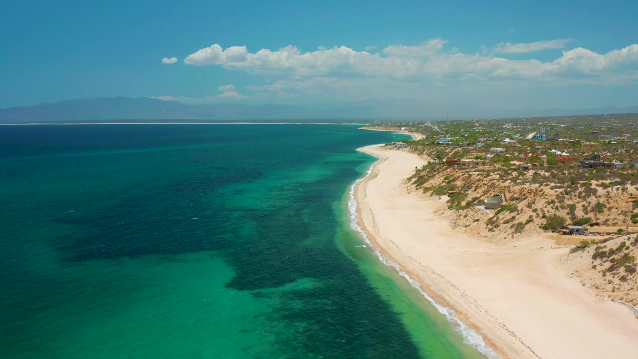 Wide aerial coastline with dark blue sea, green shallows, sand, and inland buildings.