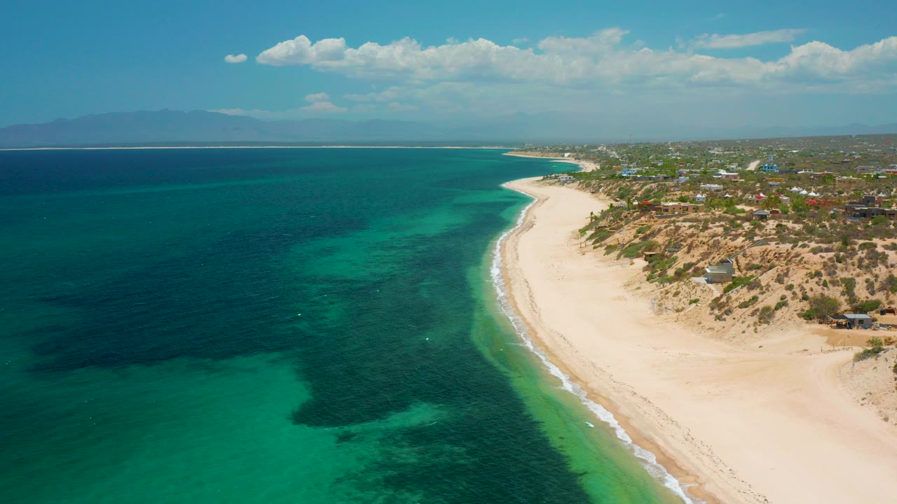 Opening aerial frame over El Sargento La Ventana, Mexico, showing turquoise and deep blue water, pale beach, inland settlement, distant mountains, scattered clouds, and no visible people.