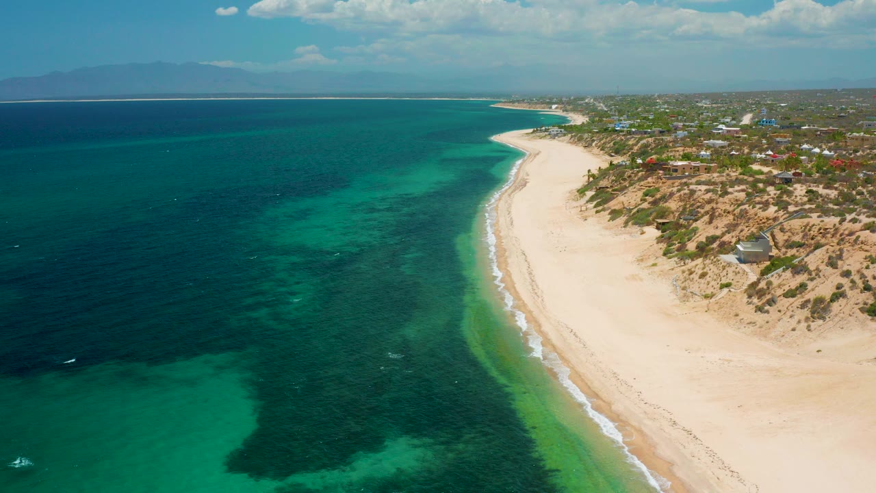 Opening aerial frame over El Sargento La Ventana, Mexico, with pale sand curving beside turquoise shallows, darker offshore water, coastal dunes, distant buildings, mountains on the horizon, and no visible people.