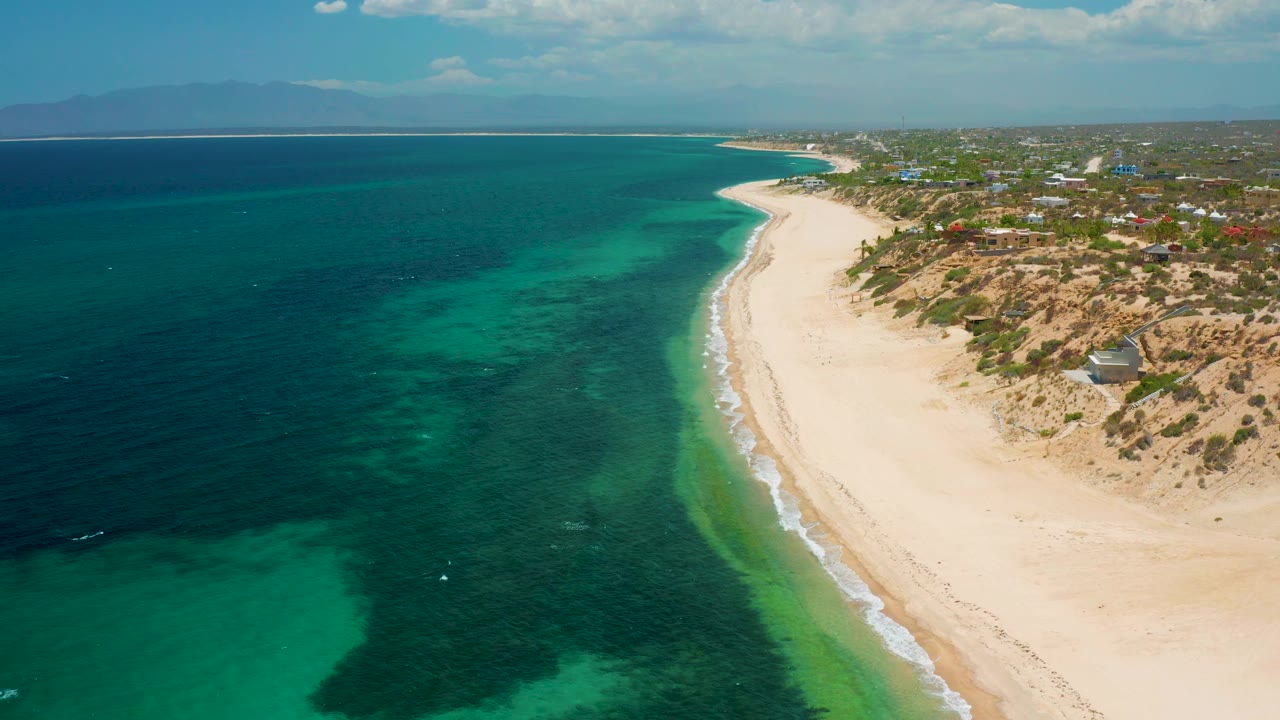 Aerial frame at 0:02 in El Sargento La Ventana, Mexico, with deeper blue water on the left, a broad turquoise band over shallow seabed, small surf along the sand, dune lots and scattered structures to the right, and no visible people.