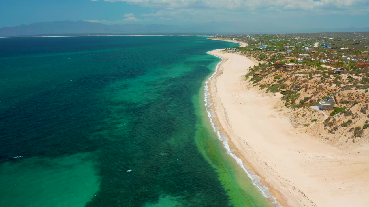 Mid-clip aerial frame over El Sargento La Ventana, Mexico, showing a wide empty beach, thin white waves, green-blue shallows, darker sea beyond, dry dunes with brush, distant rooftops, and no visible people.