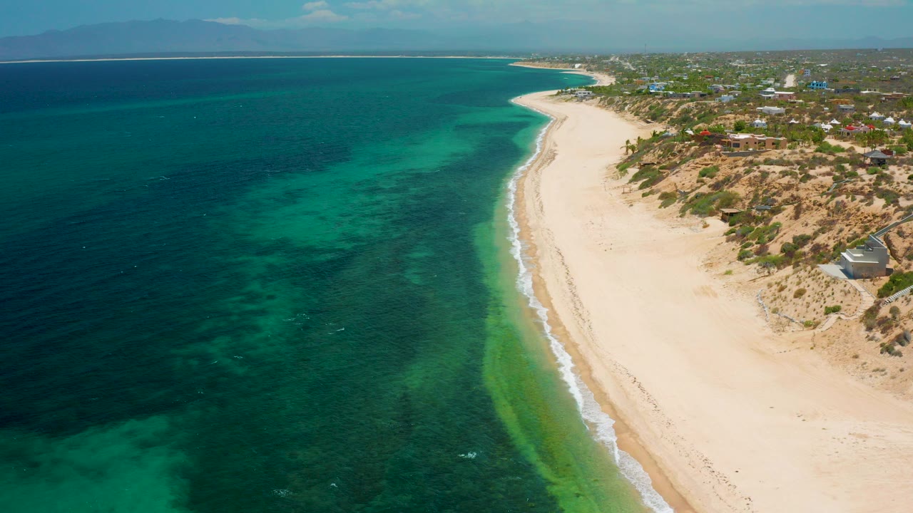 High aerial frame over El Sargento La Ventana, Mexico, showing dark blue ocean on the left, a pale tan beach curving into the distance, green shallow-water algae bands, scrubby dunes, inland buildings, and no visible people.