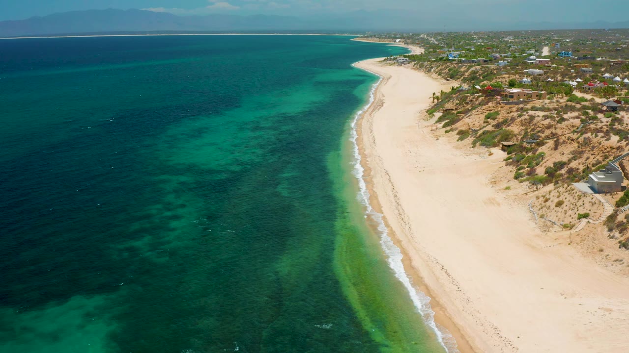 Drone frame over El Sargento La Ventana, Mexico, showing the coastline curving toward the horizon, turquoise-green shallows beside dark blue water, a pale beach strip, low dunes, scattered structures inland, and no visible people.
