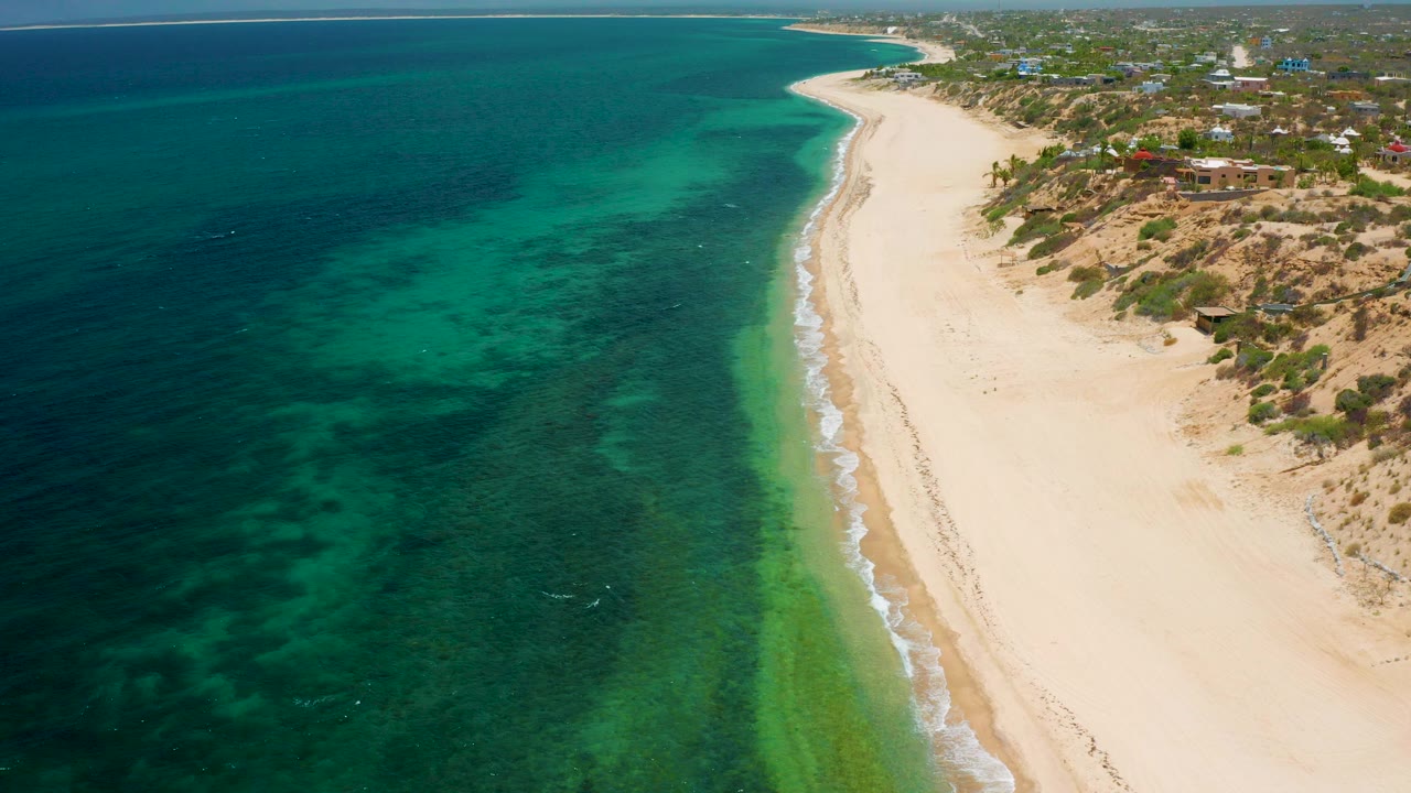 Aerial frame over El Sargento La Ventana, Mexico showing turquoise water on the left, a pale sandy beach curving along the center, low buildings on the dunes to the right, and no visible people.