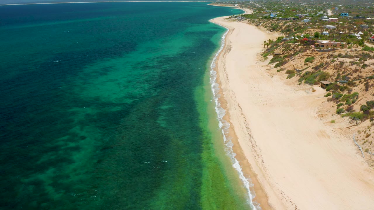 High aerial frame over El Sargento La Ventana, Mexico showing a long diagonal surf line, clear turquoise patches in the ocean, pale sand on the right, coastal buildings beyond the dunes, and no visible people.
