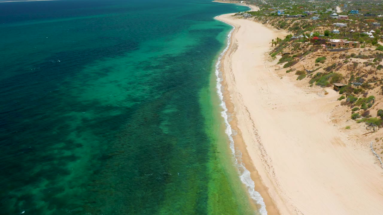 Aerial coastal view with teal water, empty sand, and dune-side buildings.