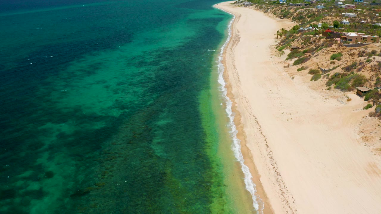 High aerial view over El Sargento La Ventana, Mexico, with dark green and turquoise water meeting a pale sandy shoreline and no visible people.
