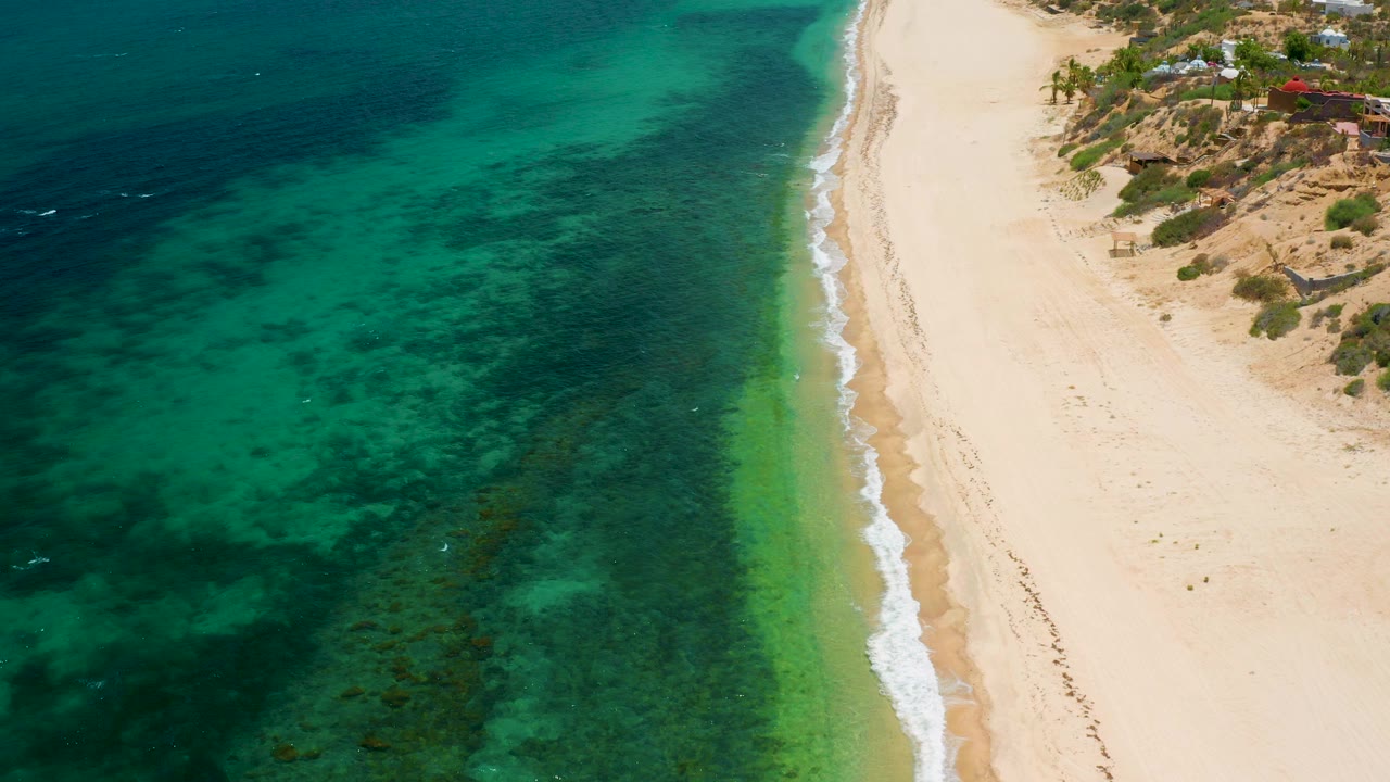 High aerial view of El Sargento La Ventana coastline with teal green ocean on the left, pale sand beach on the right, small shore break, and no visible people.