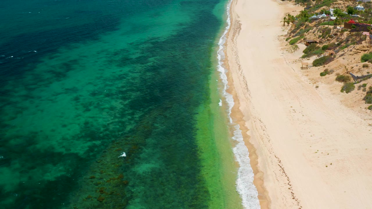 Overhead El Sargento La Ventana coast frame showing teal ocean with mottled dark patches, a thin green shallows band near the sand, dry beach, and no visible people.