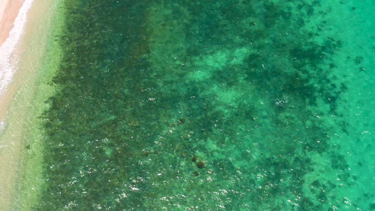 Aerial-looking downward frame from El Sargento La Ventana, Mexico showing the beach margin on the left, darker reef texture through clear water, and no visible people.