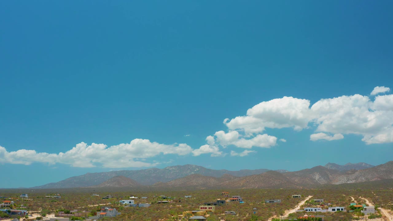 Frame at 0:02 keeps the El Sargento La Ventana horizon centered, with blue sky, puffy clouds, brown mountains, dusty roads, low buildings, and no visible people.