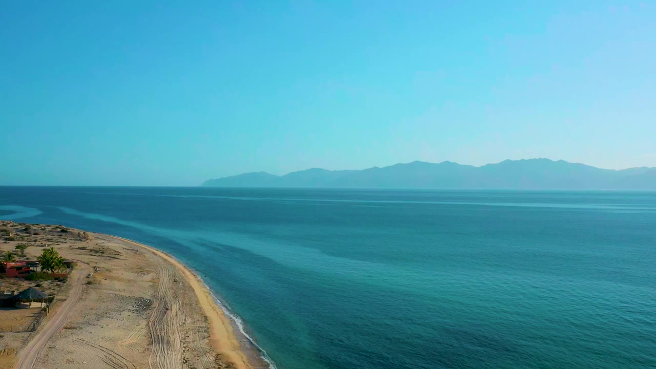 Opening aerial frame over El Sargento La Ventana, Mexico, with sandy coast on the left, blue-green ocean on the right, distant mountains on the horizon, and no visible people.
