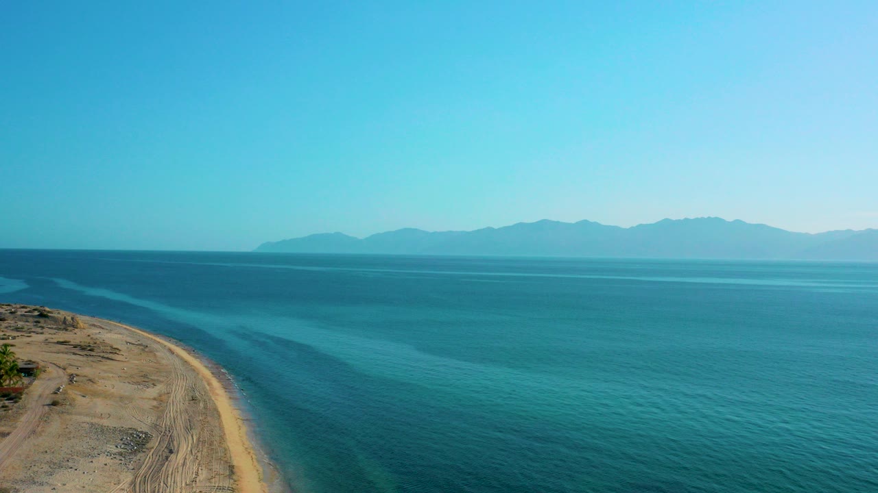 Opening aerial frame over El Sargento La Ventana, Mexico, with sandy shore at left, turquoise shallows, deeper blue water, distant mountains, and no visible people.