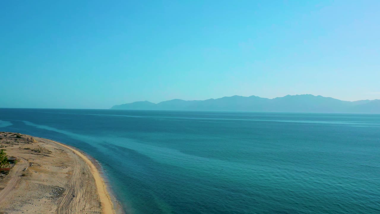 A high view in El Sargento La Ventana, Mexico, shows tan beach at left, layered blue water across the frame, faint surface texture, mountain horizon, and no visible people.