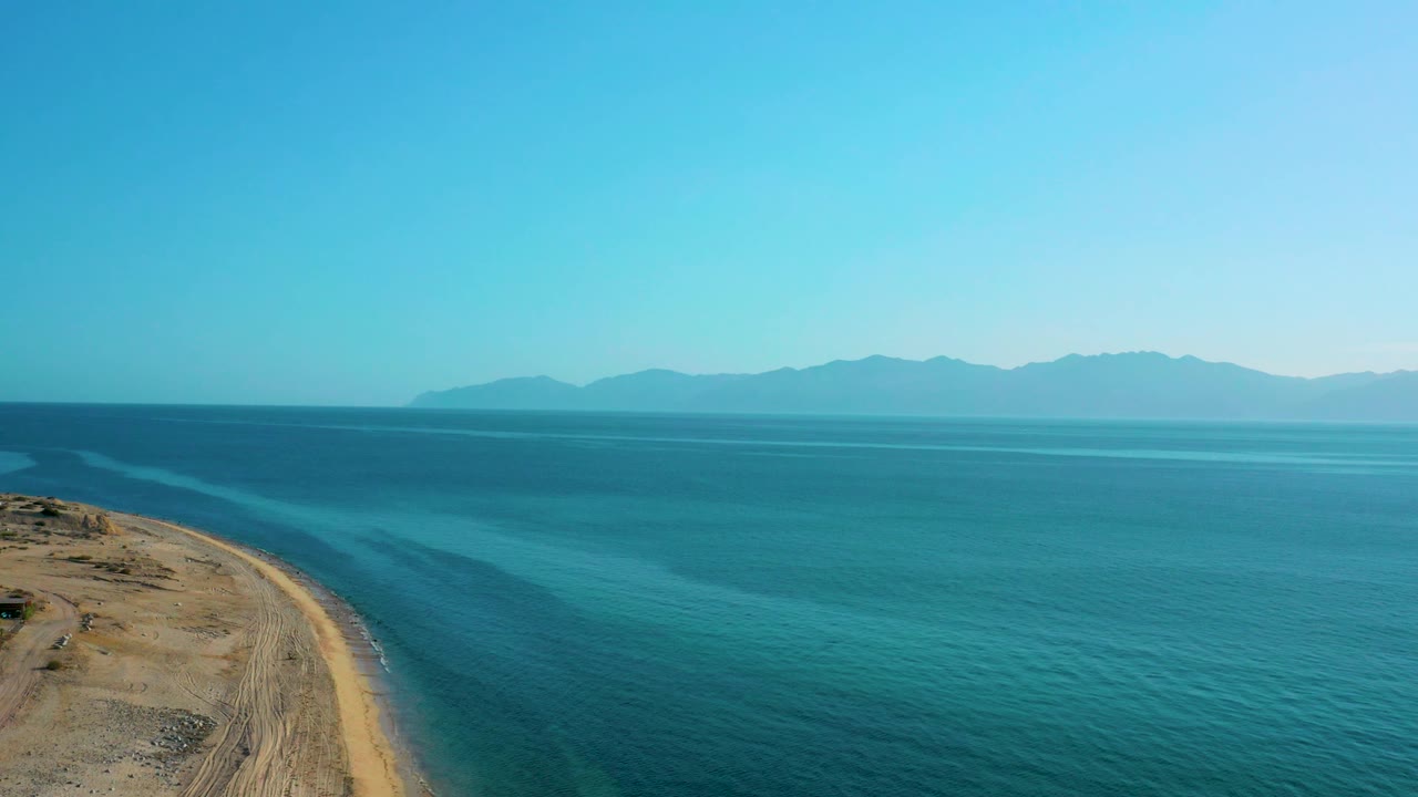 El Sargento La Ventana coastline seen from above, with a narrow sandy edge at left, broad turquoise and dark blue sea, hazy mountains in the distance, and no visible people.