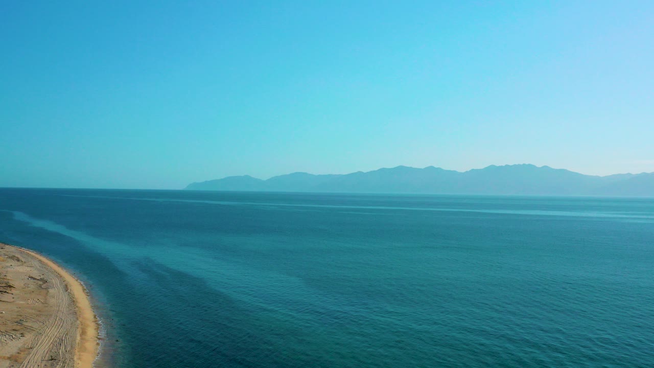 Opening frame of El Sargento La Ventana, Mexico with blue ocean across the frame, a sandy shoreline along the left edge, distant mountains on the horizon, and no visible people.