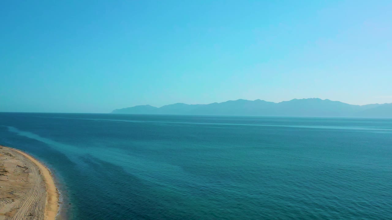 El Sargento La Ventana shoreline remains at the left as turquoise water spreads toward distant mountains under a bright sky, with no visible people.