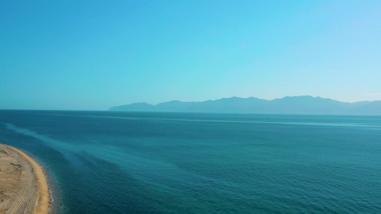 A sandy point on the left edge meets darker and lighter bands of blue water, with low mountains across the horizon in El Sargento La Ventana and no visible people.
